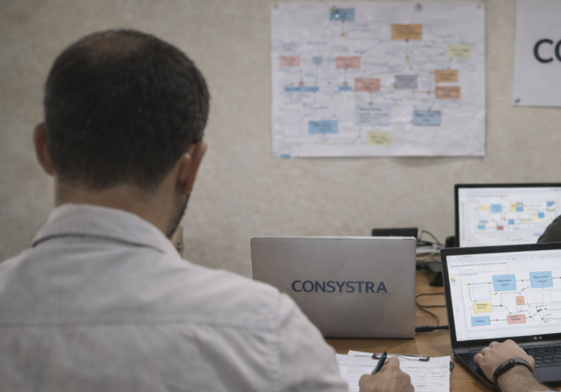 A person working at a desk with two laptops displaying diagrams, and a wall behind with a flowchart and a sign that says 'CONSYSTRA'.